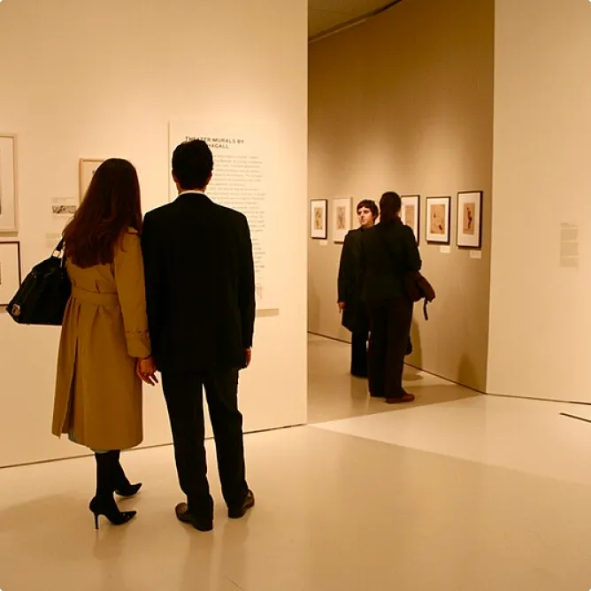 Visitors view framed artworks on the walls of a softly lit museum gallery. A couple stands in the foreground reading wall text, while others observe artworks in the background.