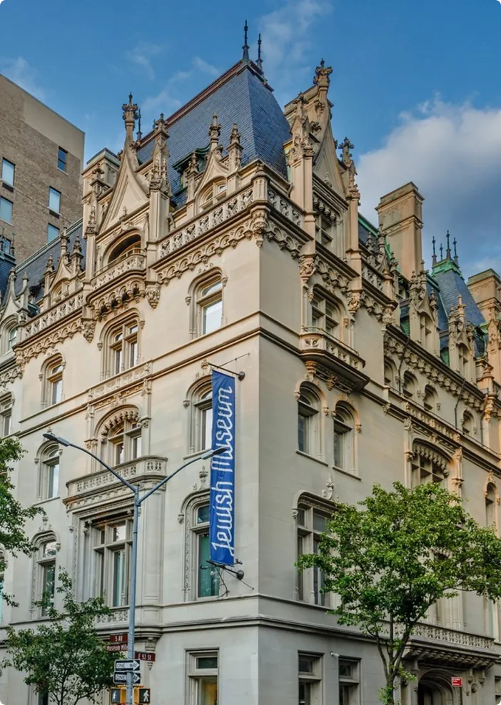 Exterior view of the Jewish Museum building with ornate stone architecture, arched windows, decorative carvings, and a blue vertical banner displaying the museum’s name.