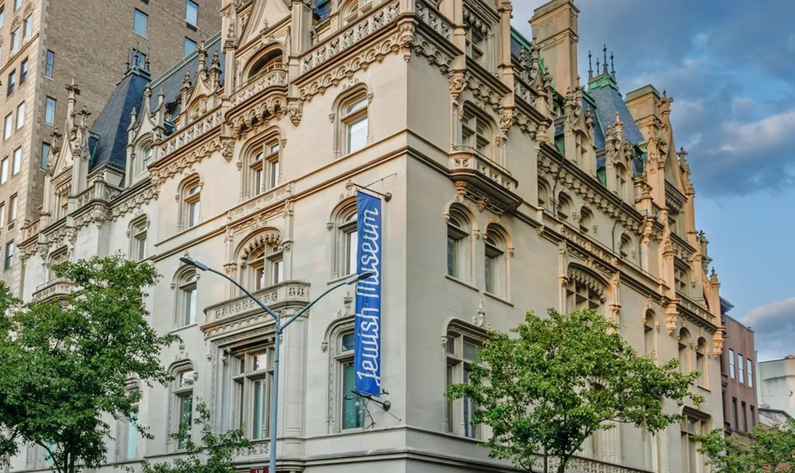 Exterior view of the Jewish Museum building with ornate stone architecture, arched windows, decorative carvings, and a blue vertical banner displaying the museum’s name.