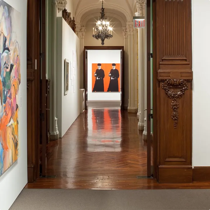 Interior view of a museum gallery with ornate wood door frames and a polished wood floor, leading to a hallway with chandeliers. Colorful abstract paintings hang on the left and right walls, and a large artwork with two black-clad figures on a red background is visible at the far end.