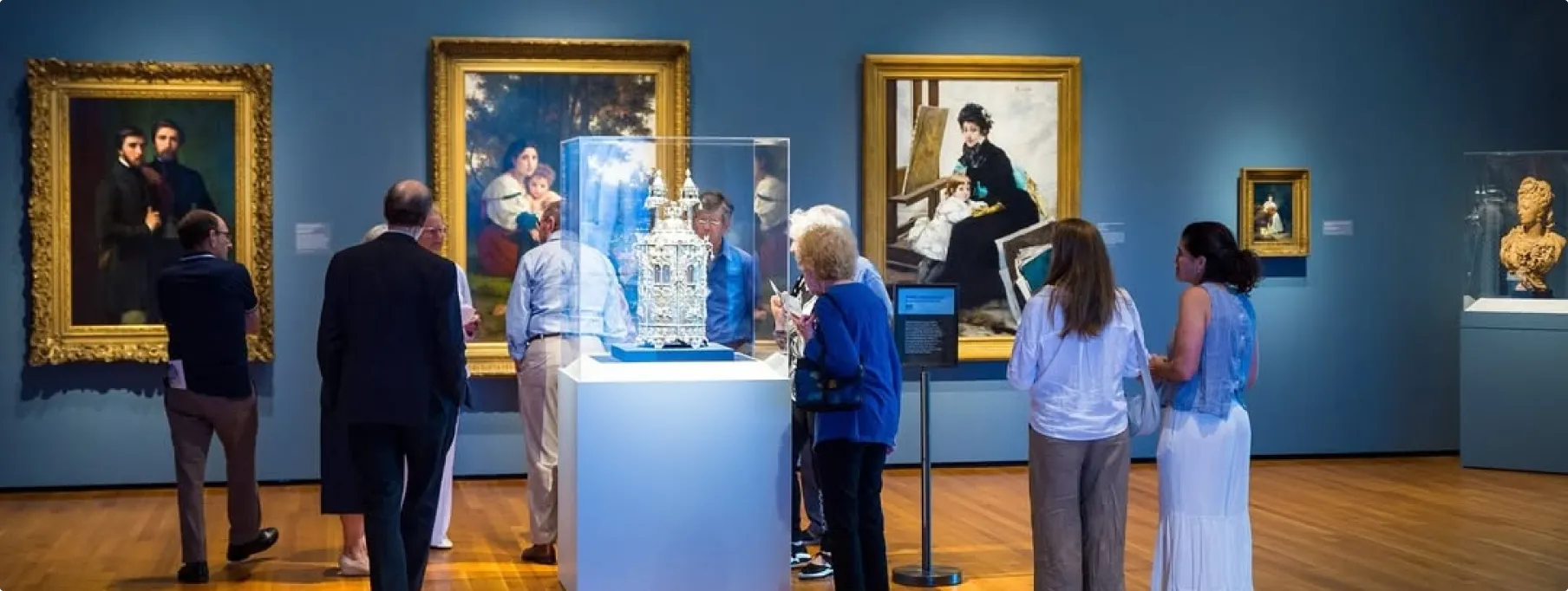 A group of museum visitors stands in front of a gallery wall painted blue, viewing a collection of 19th-century paintings in ornate gold frames. In the center, a large ornate silver ceremonial object is displayed in a glass case on a pedestal. Visitors are closely observing the exhibit, reading labels, and discussing the artwork.