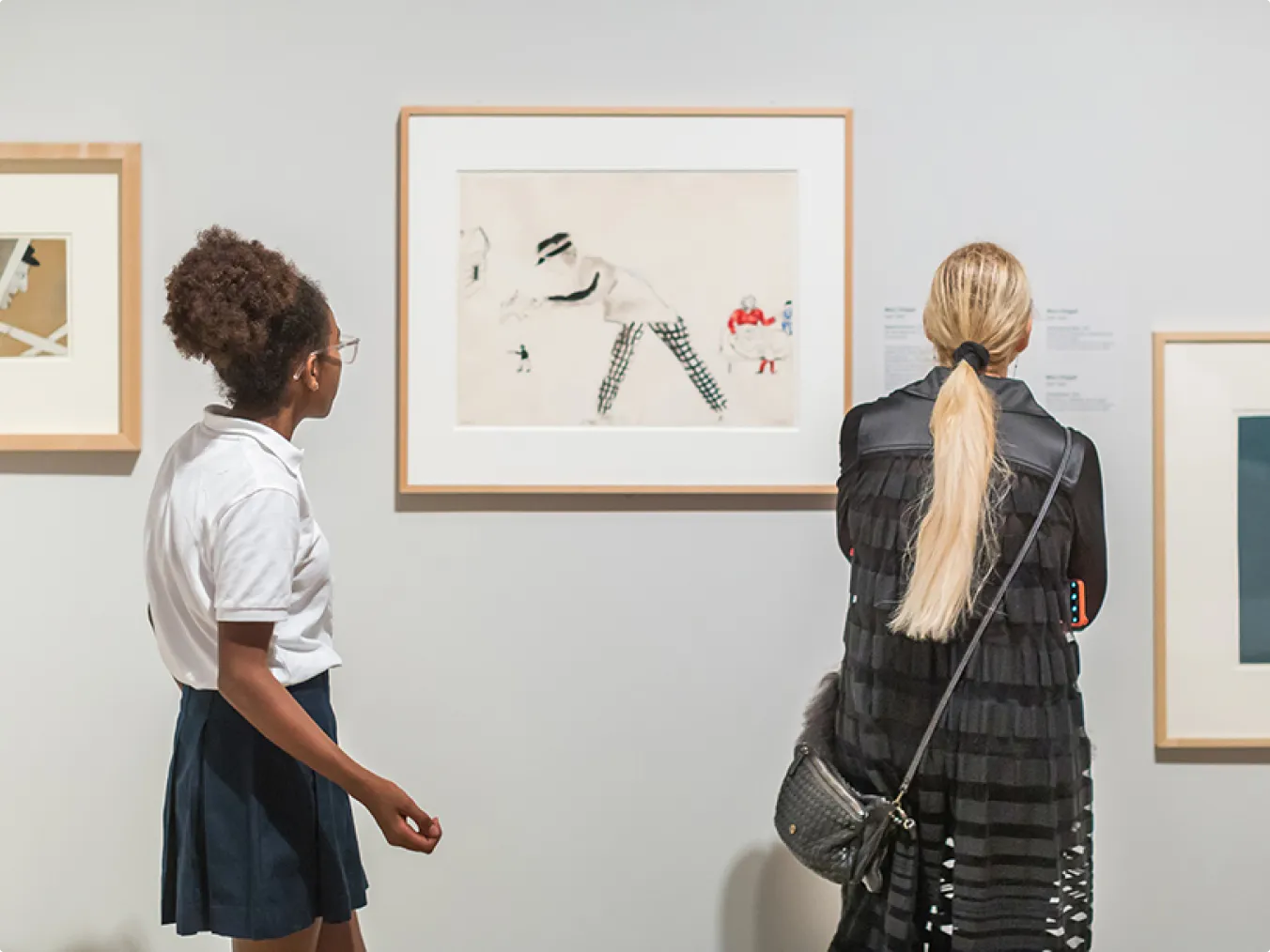 Two visitors view framed artworks on a gallery wall. The person on the left wears a white shirt and dark skirt; the person on the right has long blonde hair tied back and wears a black outfit with horizontal stripes, carrying a black crossbody purse.