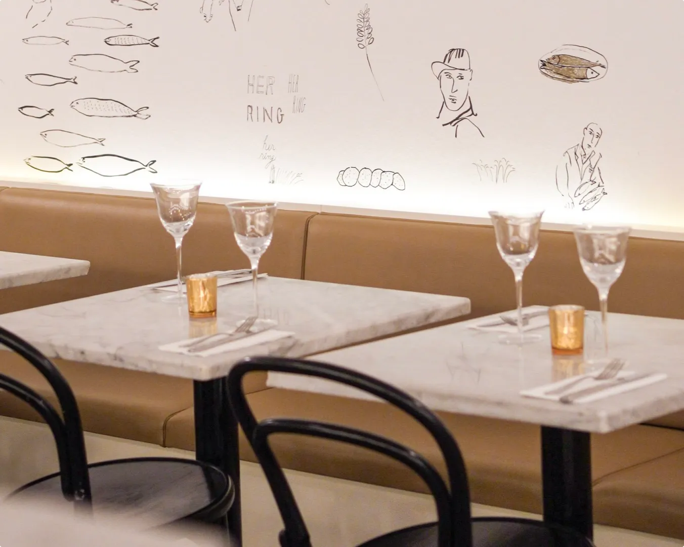 Close-up of restaurant tables with marble tops, set with glassware, utensils, and candles; illustrated wall art in the background.