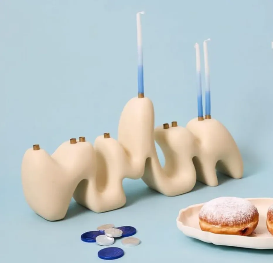 Modern cream-colored Hanukkah menorah with organic, sculptural shapes, three blue-and-white candles, chocolate gelt coins, and powdered jelly donuts on a plate, set against a light blue background.