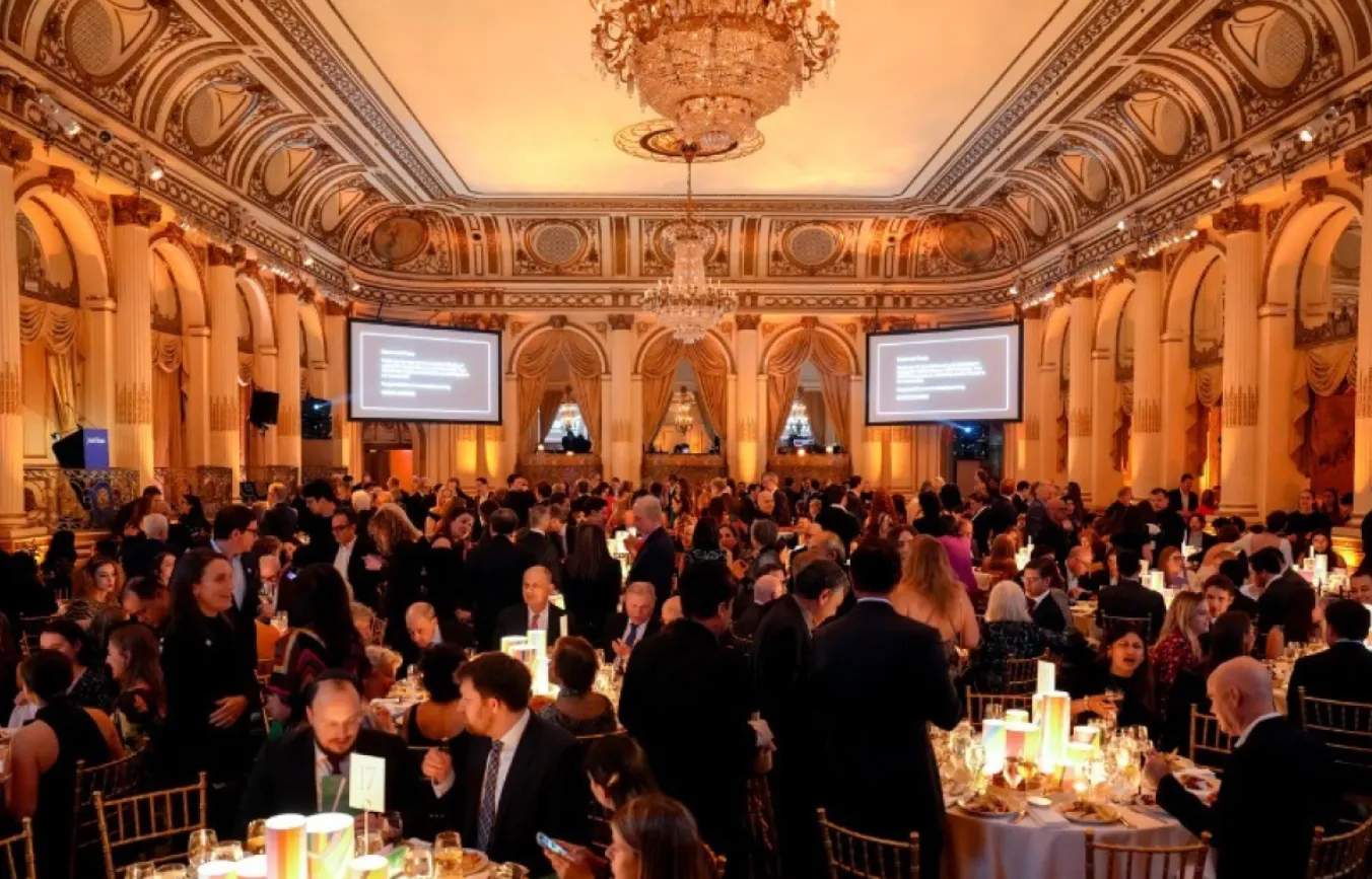 Guests seated at round tables and standing in a large ballroom during a formal event with chandeliers and projection screens.