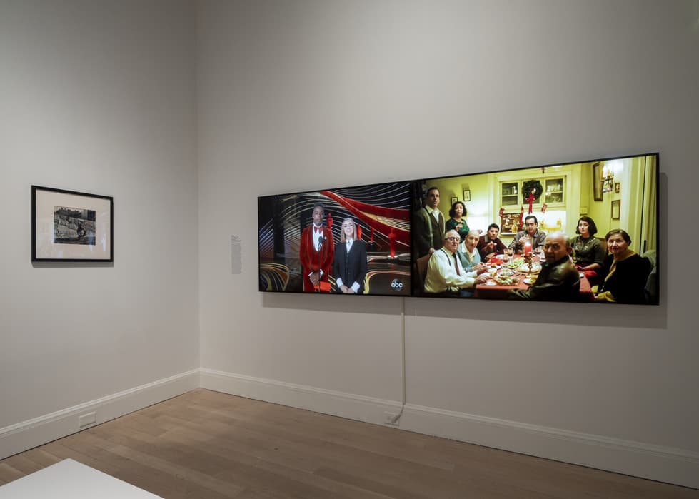 Gallery wall displaying a small framed black-and-white photograph on the left and two large horizontal video screens on the right; the left video screen shows two people standing on an awards stage, while the right screen shows a group of people seated around a dining table in a warmly lit home interior.