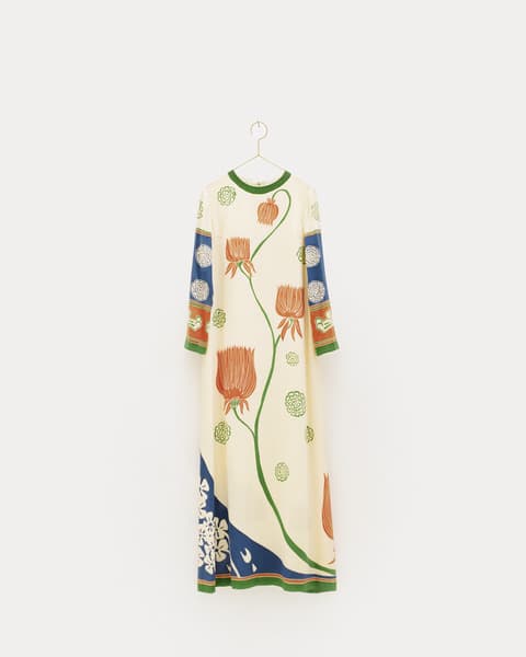 Long cream-colored dress with botanical designs of large orange flowers and green stems, featuring blue and green patterned accents on the sleeves and hem, displayed on a hanger against a white background.
