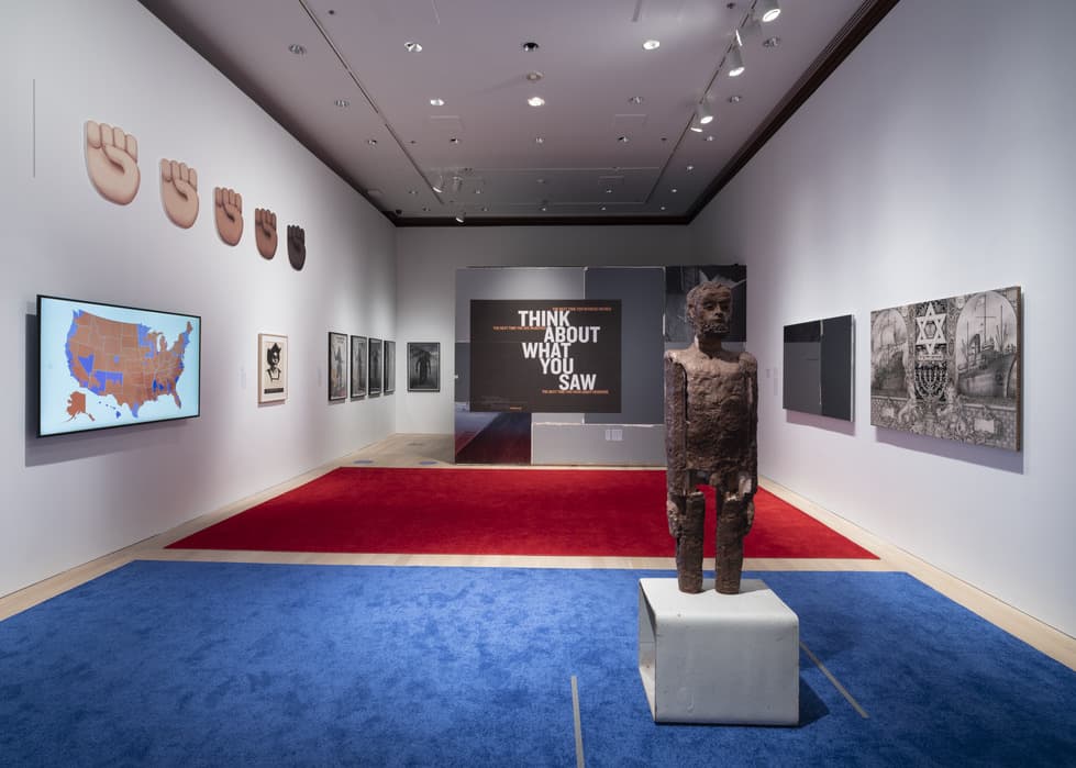 Gallery installation with a red and blue carpeted floor, featuring a central wooden sculpture of a standing human figure. On the left wall, there are raised fist symbols in multiple skin tones, a digital map of the United States, and framed artworks. On the right wall are monochrome drawings and prints, including a detailed illustration with a Star of David. The far wall displays bold text reading “THINK ABOUT WHAT YOU SAW.”