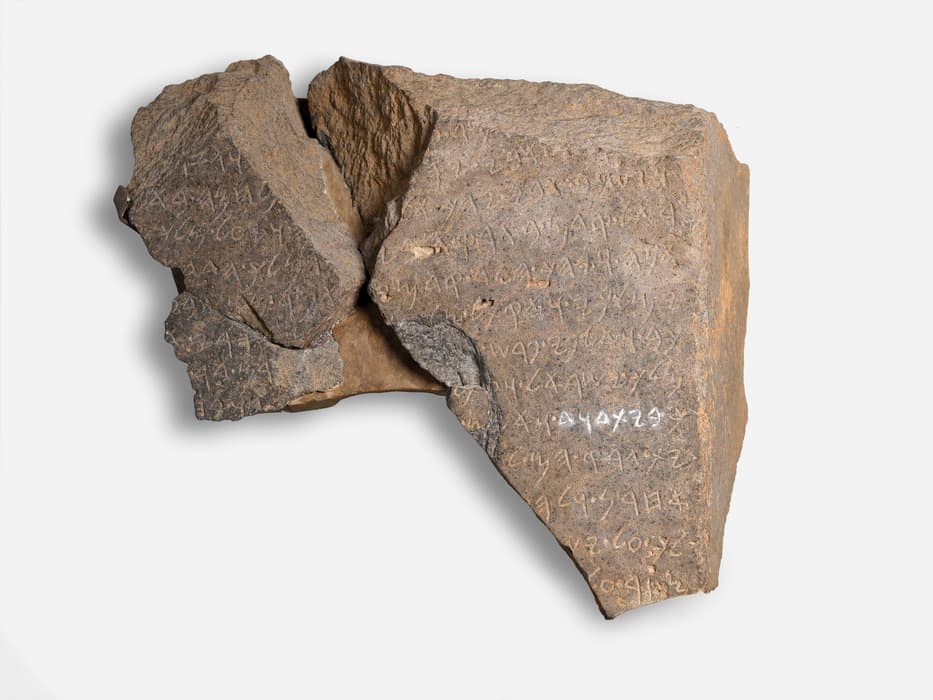 Ancient stone fragment inscribed with lines of text in an early script, showing multiple breaks and rough edges, displayed against a white background.
