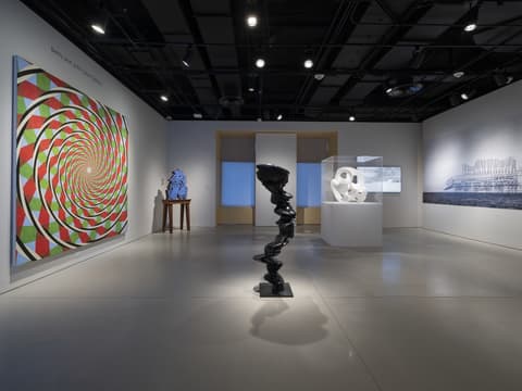 A modern art gallery installation featuring various abstract works. On the left wall hangs a large, colorful spiral painting with concentric circles in red, green, black, and white, creating an optical illusion effect. In the center foreground stands a tall, glossy black abstract sculpture on a pedestal, composed of a twisting, organic form. Toward the back, on a wooden table, sits a blue abstract sculpture, while a white translucent geometric sculpture is displayed in a glass case in the center rear. The right wall features a large grayscale photograph or mural of an architectural structure. The gallery space has a polished gray floor, dark ceiling with track lighting, and minimalist white walls.