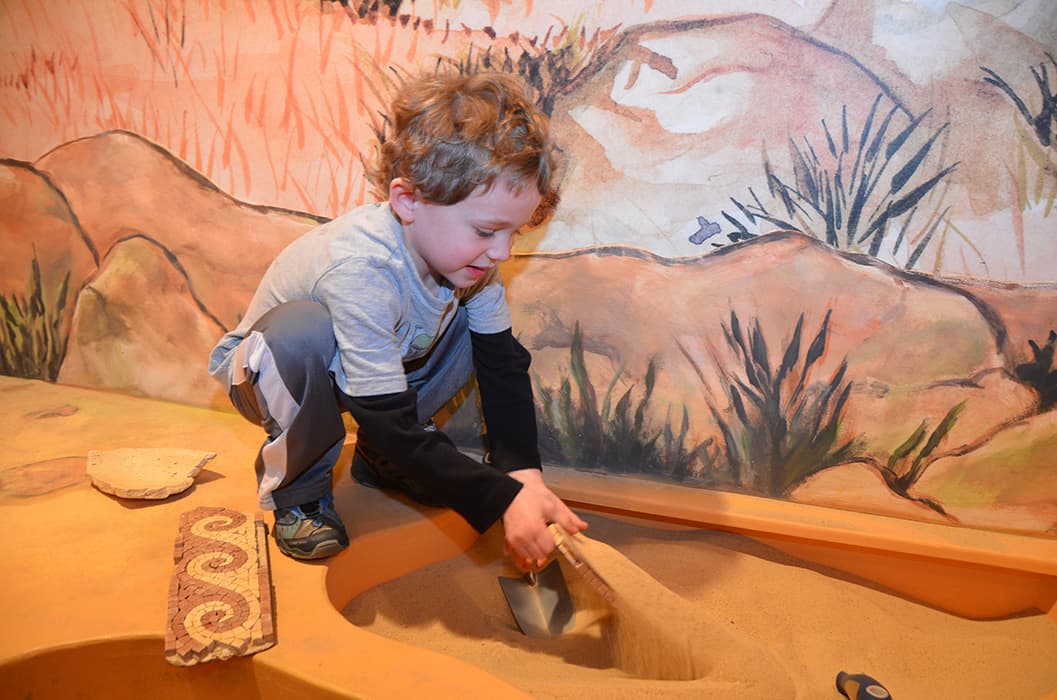 A young boy crouches in a simulated dig site, using a brush to sweep sand off a buried object. Fragmented artifacts lie nearby, with a desert-themed mural in the background.