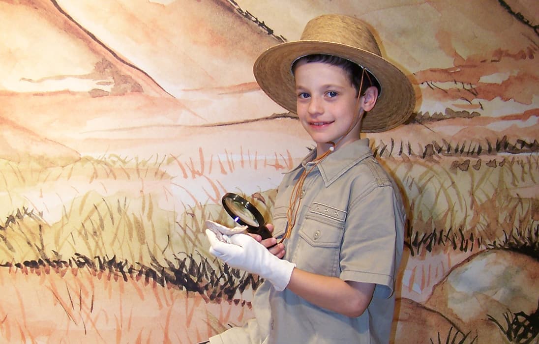 A young boy dressed as an archaeologist holds a magnifying glass and an artifact while wearing white gloves and a wide-brimmed straw hat. He stands in front of a painted desert landscape backdrop.