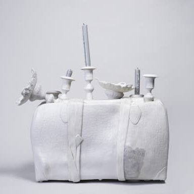 Sculpture of a white suitcase wrapped with straps, containing broken candlesticks, plates, and other domestic objects protruding from the top.