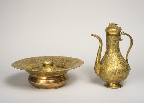 Ornate gold-colored ritual washing set consisting of a wide, shallow basin with raised decorative edges and a tall ewer with a long curved spout, slender neck, and engraved floral patterns on both pieces.