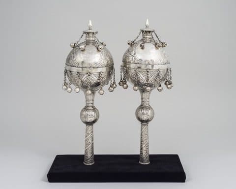 Pair of silver Torah finials with spherical tops, hanging bells, and intricate engraved patterns, displayed upright on a black base against a light gray background.