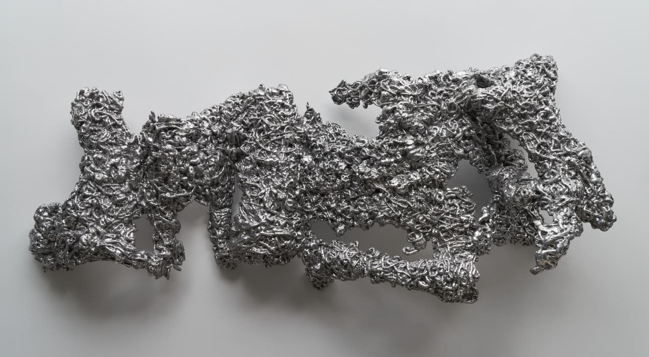 An abstract metallic sculpture mounted on a light gray background. The irregular, organic form appears as a tangled mass of shiny, silver-colored material with numerous crevices, folds, and protrusions. The reflective surface highlights the intricate textures and complex layering, giving it a fluid, almost molten appearance.
