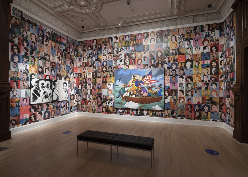 Gallery room with walls covered in colorful celebrity portraits; large central painting depicting George Washington Carver Crossing the Delaware; two black and white portraits mounted to the left; black bench centered on wooden floor beneath the artworks.