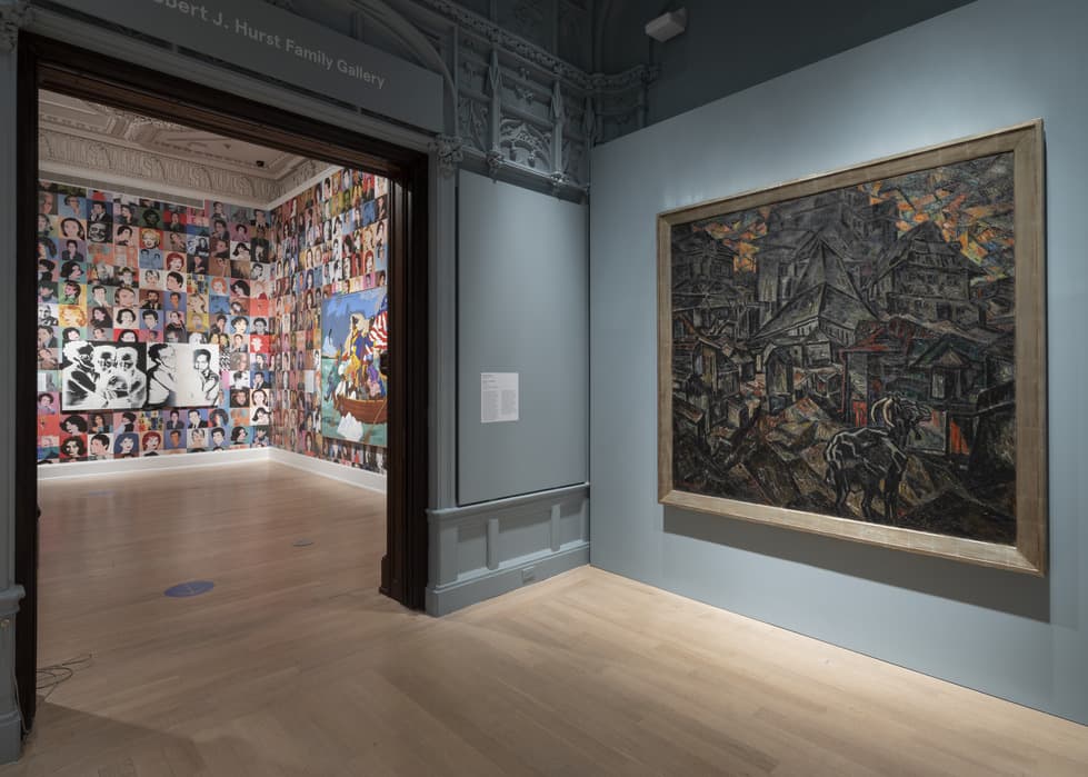 Gallery view showing a dark-toned painting of clustered buildings and a horse on the right wall; doorway leads into adjacent room with walls covered in colorful celebrity portraits and a large painting of George Washington Carver Crossing the Delaware.