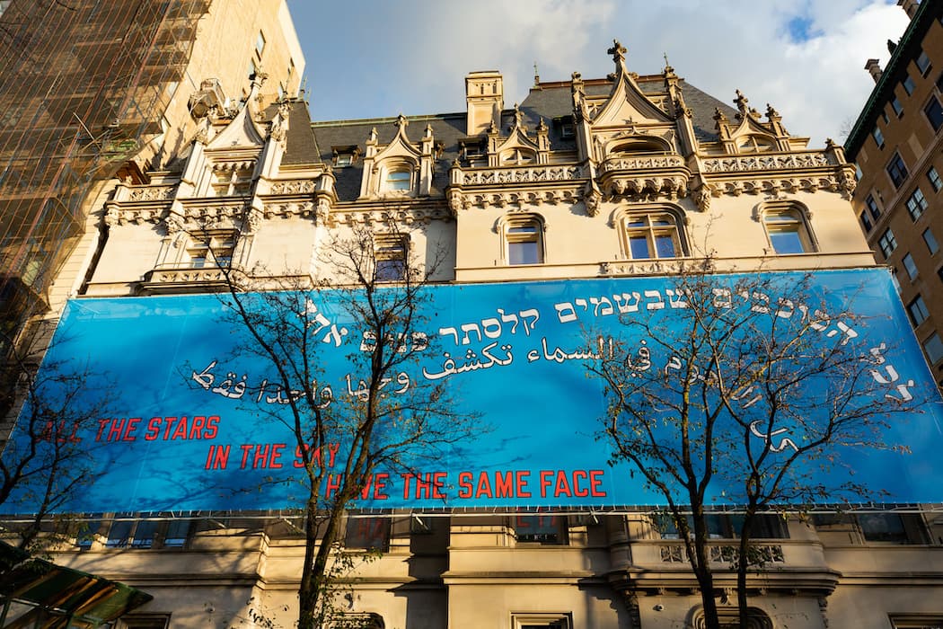Historic stone building with ornate detailing and a large blue banner stretched across the facade. The banner displays text in Hebrew, Arabic, and English, reading