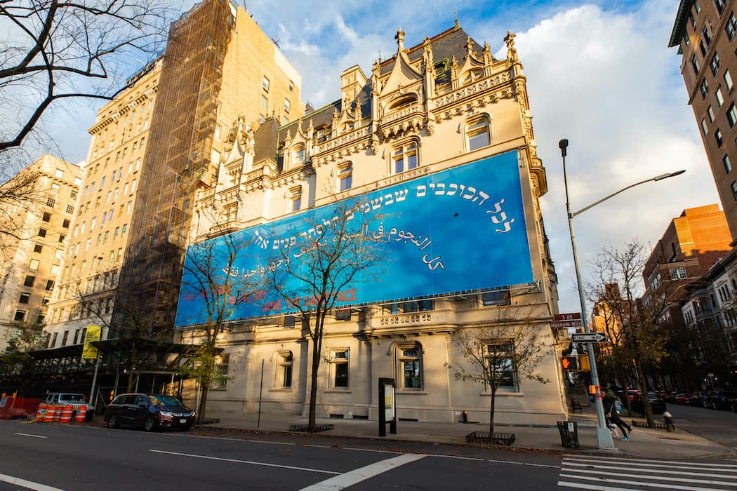 Exterior of a historic stone building with ornate architectural details and a large blue banner covering its facade. The banner features text in multiple languages including Hebrew, Arabic, and English, reading “ALL THE STARS IN THE SKY HAVE THE SAME FACE.” The building sits on a city street corner with trees and surrounding tall buildings.