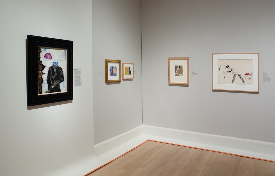 Gallery corner featuring a variety of framed modernist artworks, including abstract compositions and figurative scenes, displayed on soft gray walls with warm wood flooring and simple wall labels.