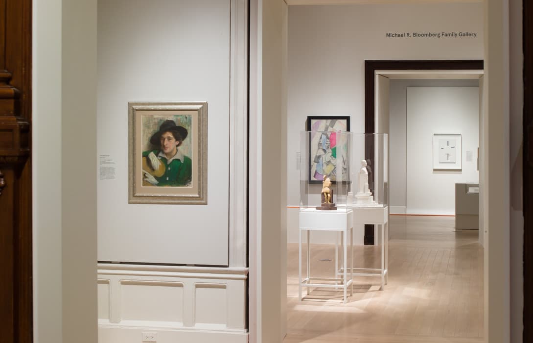 View through multiple gallery doorways showing a portrait of a woman in green, modernist paintings, and two small sculptures in clear display cases under soft lighting.