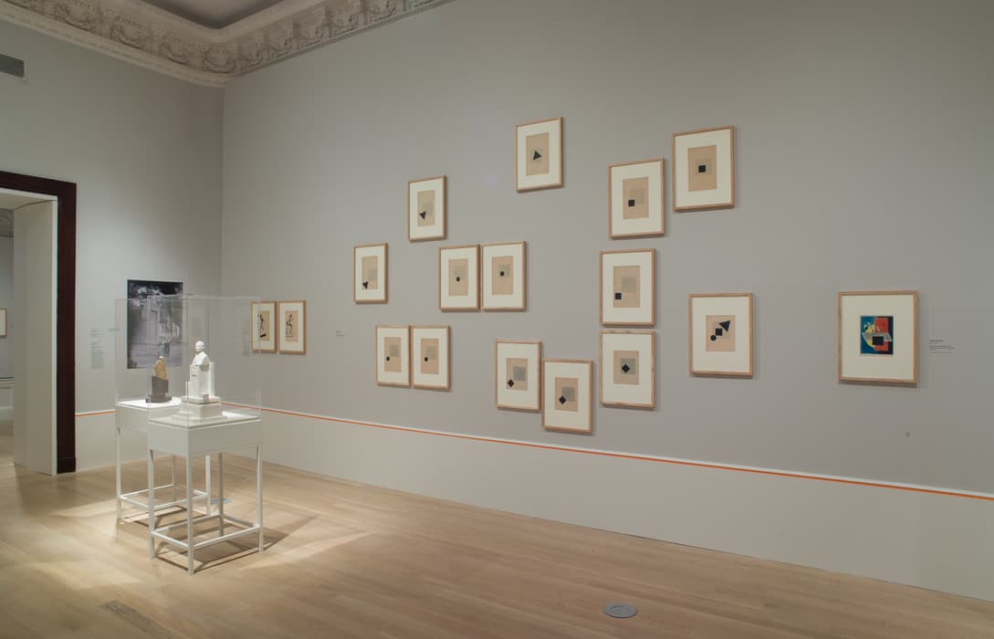 Gallery wall displaying a geometric arrangement of framed abstract prints, mostly featuring black shapes on neutral backgrounds, alongside a colorful modernist work; two small sculptures are shown in a glass case on white pedestals in the foreground.