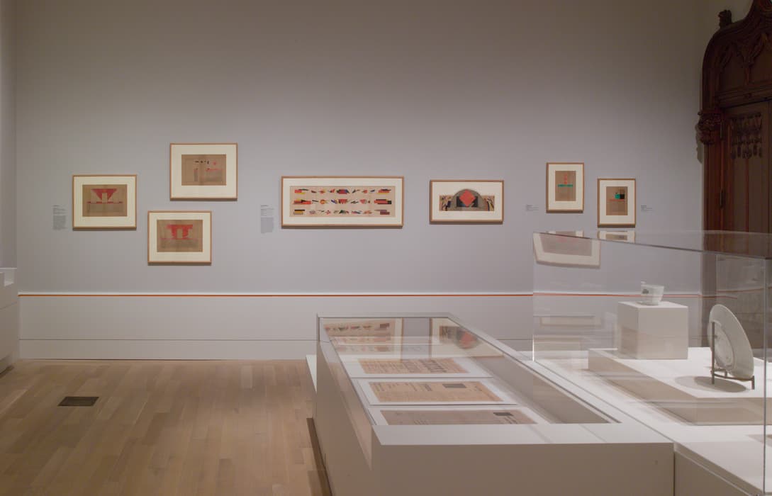 Museum gallery featuring framed abstract works with geometric forms in red, orange, and green on a gray wall; display cases in the foreground hold documents and ceramic pieces.