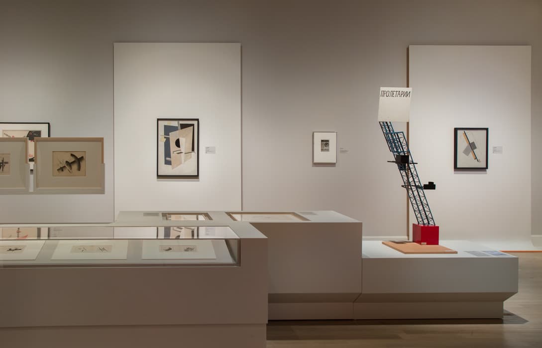Exhibition gallery displaying Constructivist artworks, including framed geometric compositions and a 3D architectural model featuring a slanted lattice tower with Russian text. Display cases in the foreground contain sketches and prints.
