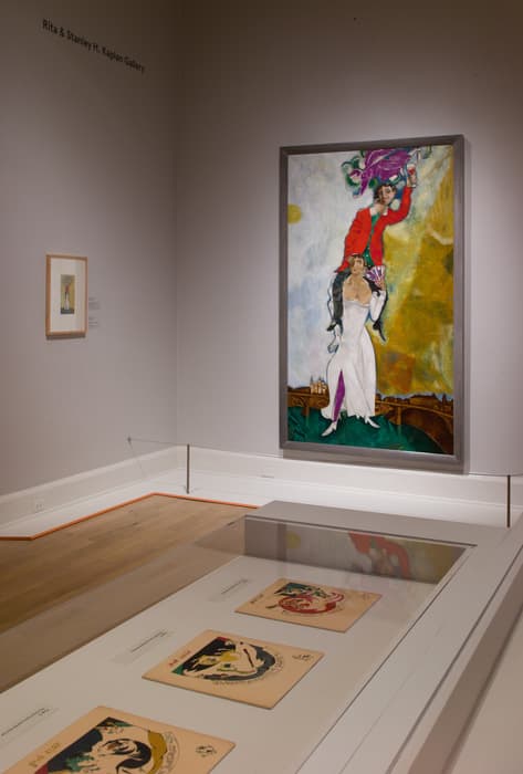 Gallery view featuring a large vertical painting of a woman in a white dress carrying a man in a red coat on her shoulders, with vibrant colors and a cityscape background. Nearby, several illustrated works are displayed in a glass case, and a smaller framed piece hangs on the adjacent wall.