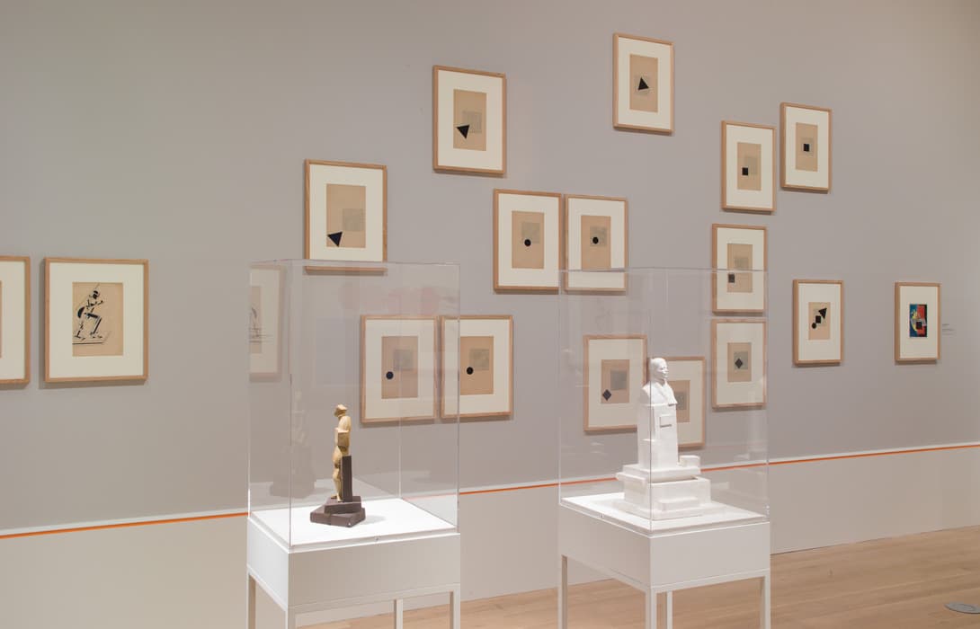 Museum gallery with abstract geometric prints arranged on a gray wall and two small modernist sculptures displayed in plexiglass cases.