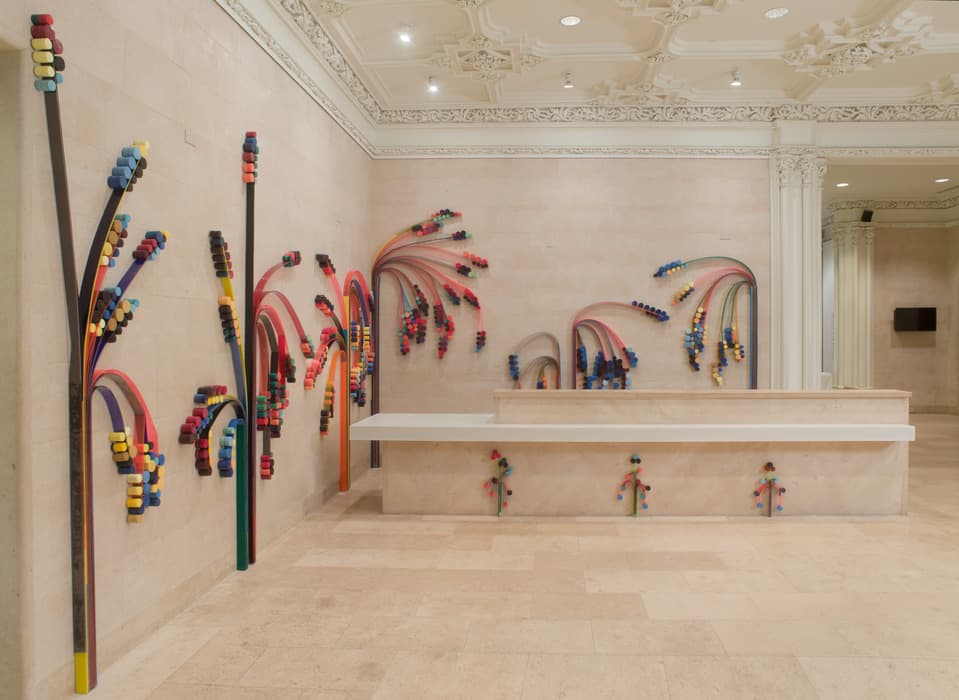 Sculptural wall installation featuring colorful cables and foam pieces arranged in palm-like arcs across a beige marble interior with ornate ceiling details.