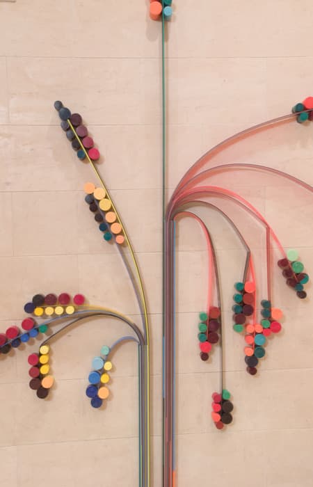 Close-up view of a sculptural wall installation composed of arcing translucent tubes and colorful foam discs arranged like stylized plant stems and blossoms, mounted on a beige tiled wall.