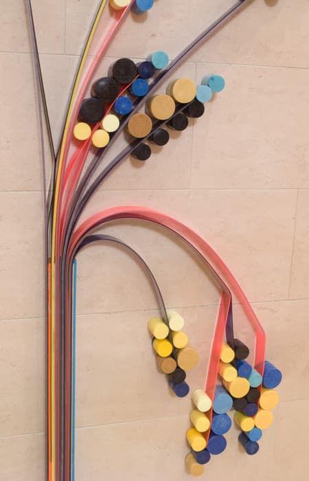 Close-up of a sculptural wall installation made of colorful foam discs attached to curved plastic tubes, resembling stylized plant stems with blooming clusters in shades of yellow, blue, black, and brown.