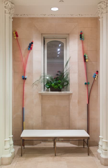 Sculptural installation featuring two tall, slender stalks made of translucent red tubing and colorful foam discs mounted on either side of a window alcove, with a bench below and a green plant on the window ledge.