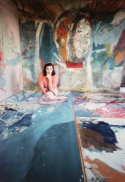 Woman sits cross-legged on the floor of a studio filled with large, colorful abstract paintings covering the floor and walls. She wears a pink blouse, light pants, and a headband, resting her chin on one hand while looking at the camera. The vibrant brushstrokes around her feature bold blues, reds, yellows, and organic shapes.