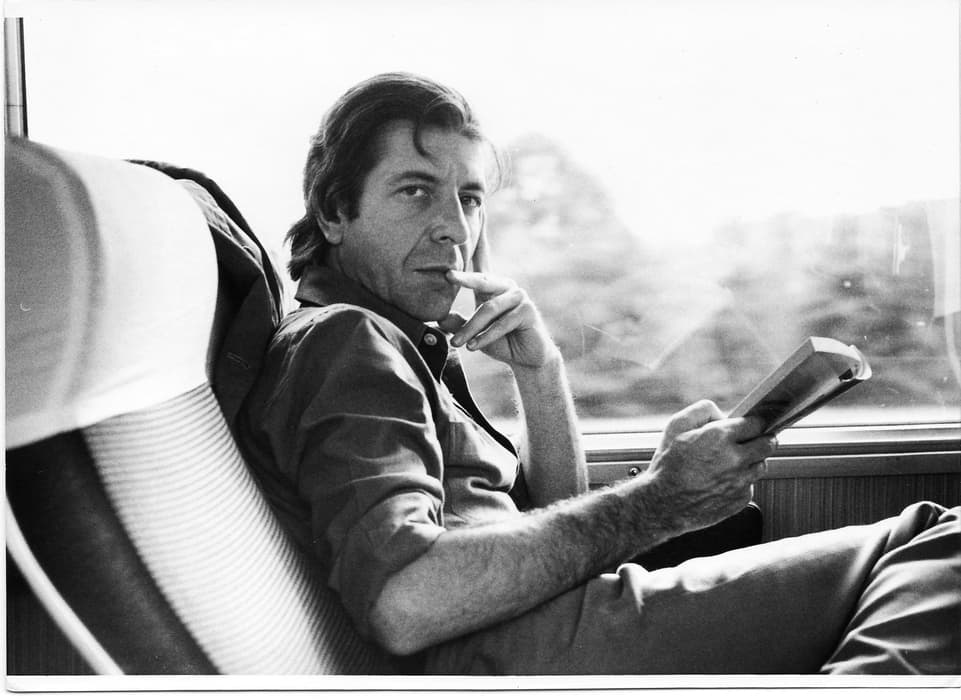 Black-and-white photo of a man sitting in a train seat, holding an open book and looking pensively toward the camera, with blurred scenery visible through the window behind him.