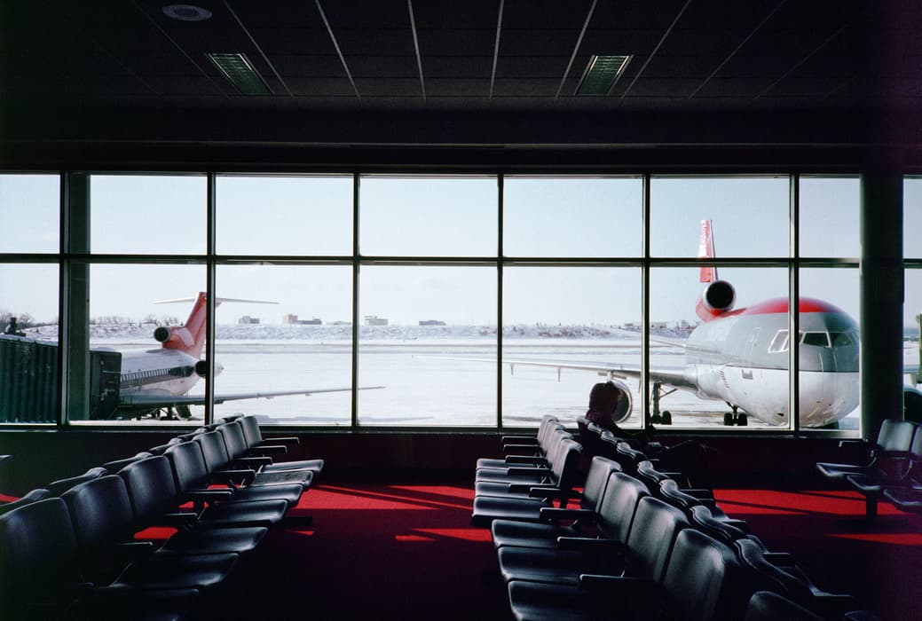 A nearly empty airport terminal with rows of black chairs on red carpet, looking out through large windows at two parked airplanes on a snow-covered tarmac; a lone passenger sits quietly near the window.