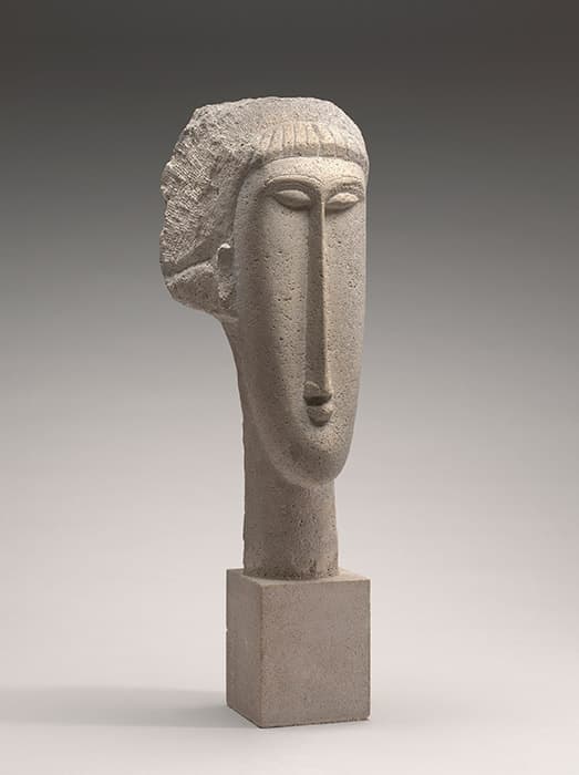 Stone sculpture of a stylized elongated head with almond-shaped eyes, a long nose, and simplified lips, mounted on a rectangular base.