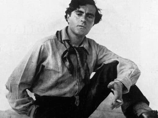 Black-and-white portrait of a man sitting on a wooden beam, wearing a loose shirt with a scarf tied at the collar, dark trousers, and boots, holding a cigarette and gazing directly at the camera.