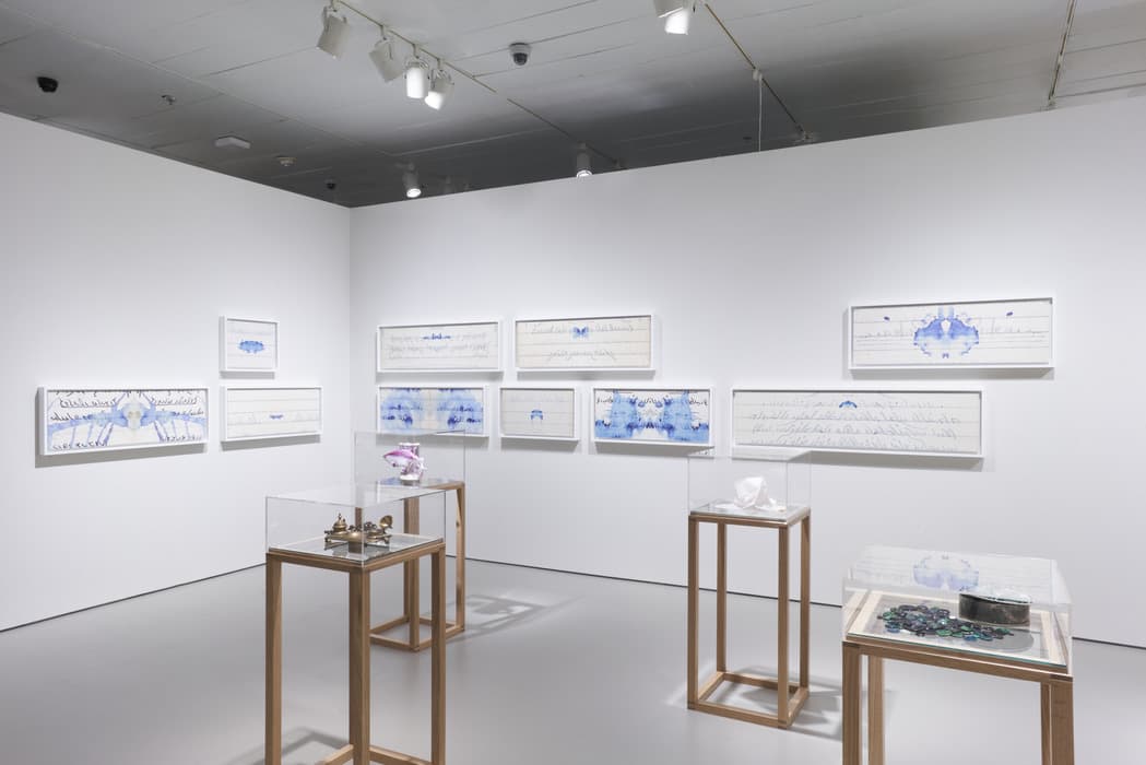 Gallery view with framed works on paper featuring blue ink blots and cursive writing on the walls, and four display cases containing small objects and sculptures on light wooden stands.