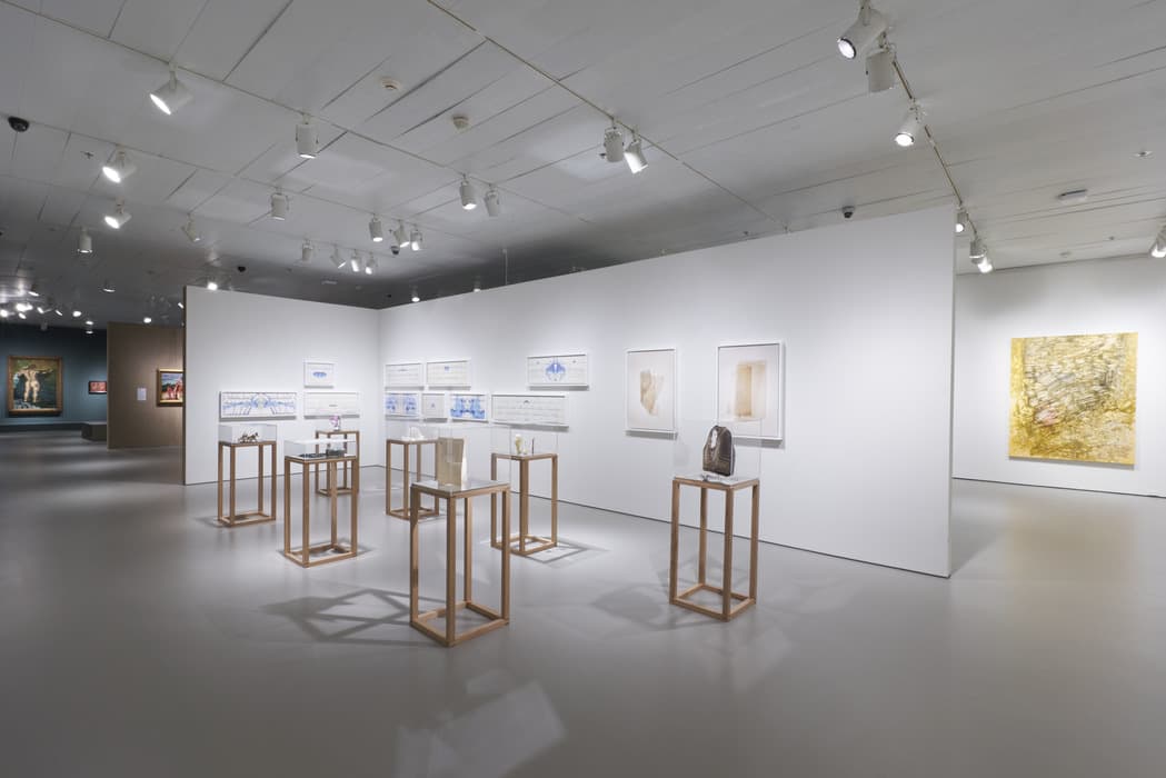 Gallery installation view featuring a group of artworks displayed on freestanding light wood pedestals with clear vitrines, containing small sculptural and artifact-like objects. Behind them, a white wall displays framed works on paper, including ink drawings and prints with abstract and calligraphic blue markings. To the right are two framed, neutral-toned prints and a small sculpture. In the background, another gallery is visible with additional paintings, including a large yellow monochrome painting on the far right and a darker-hued gallery on the left.