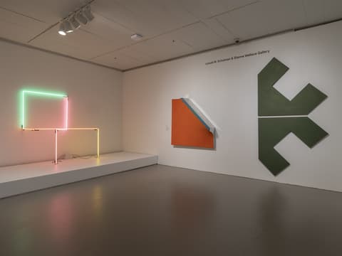 Art gallery interior with three minimalist works displayed. On the left, a neon light sculpture composed of bright green, pink, and orange horizontal and vertical lines sits on a white platform. On the center wall, a square canvas painted orange with a white and light blue diagonal stripe appears to lean forward from the wall. On the right wall, a large geometric green painting features two stacked arrow-like shapes pointing in opposite directions. The gallery has neutral lighting and gray flooring, with the label 