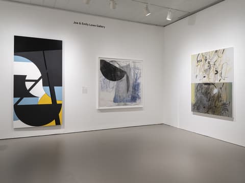 Interior view of the Joe & Emily Lowe Gallery with three contemporary abstract paintings displayed. On the left wall hangs a tall vertical painting with bold black curved and angular shapes layered over pale blue, white, and yellow geometric areas. The center features a large drawing with a translucent appearance, combining black, gray, and blue shapes, including a prominent black half-circle. On the right wall, two stacked abstract works with loose black and yellow gestural lines and smudges fill a white background, evoking a more spontaneous and organic feel.