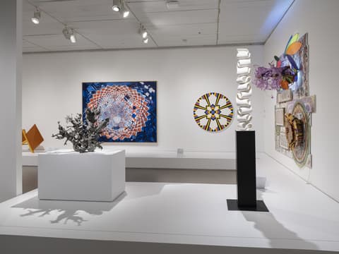 Gallery view of a contemporary art exhibit with multiple works. Left: angular orange metal sculpture; center: organic silver metal sculpture on white pedestal; back wall: geometric paintings with circular, radial designs in blue, red, and yellow; right: colorful wall-mounted mixed media assemblage with flowers, collage, and transparent shapes; vertical white sculpture on black pedestal in foreground.