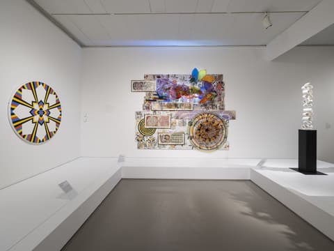 Gallery view with three contemporary artworks: left, a circular geometric painting with radiating yellow, blue, black, and white shapes; center, a large colorful mixed media wall assemblage with floral patterns, collage, and translucent forms; right, a tall white sculptural column on a black pedestal.