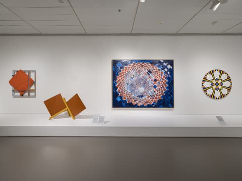 Gallery wall with four artworks: left, a red square relief sculpture; next, an angular orange geometric sculpture; center, a large blue and red mandala-like painting; right, a circular abstract painting with yellow, black, and white segments.