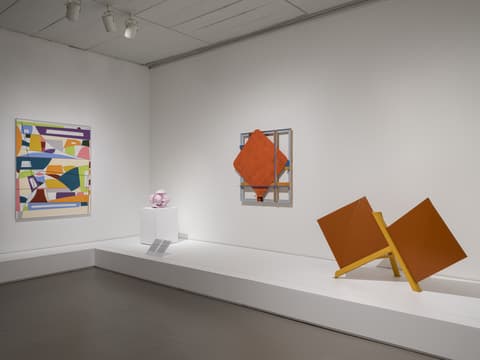 Gallery with three wall artworks and one sculpture: left, a colorful geometric abstract painting; center, a small pink abstract sculpture on a pedestal and a red diamond-shaped relief; right, a large orange angular geometric sculpture.