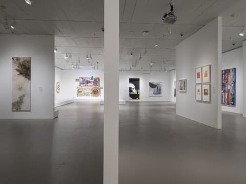 View of multiple gallery rooms separated by walls; visible are abstract paintings, colorful wall collages, framed works on paper, and sculptures.