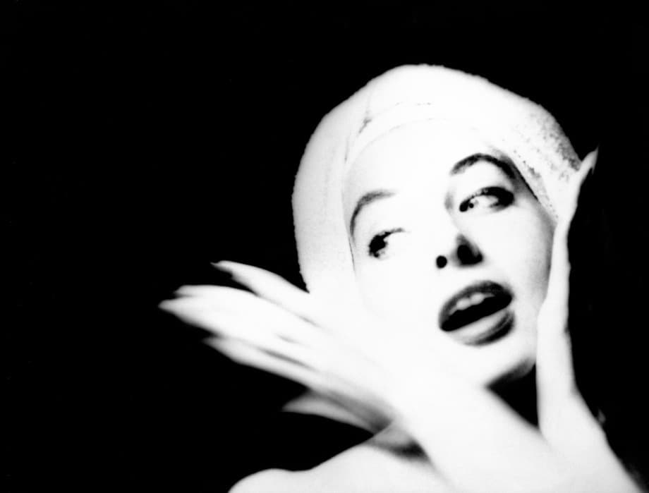 High-contrast black-and-white portrait of a woman with a towel wrapped around her head. She gazes to the side with an expressive open-mouthed smile and wide eyes. One hand rests near her face while blurred motion suggests movement in the foreground. The background is solid black, emphasizing her illuminated face.